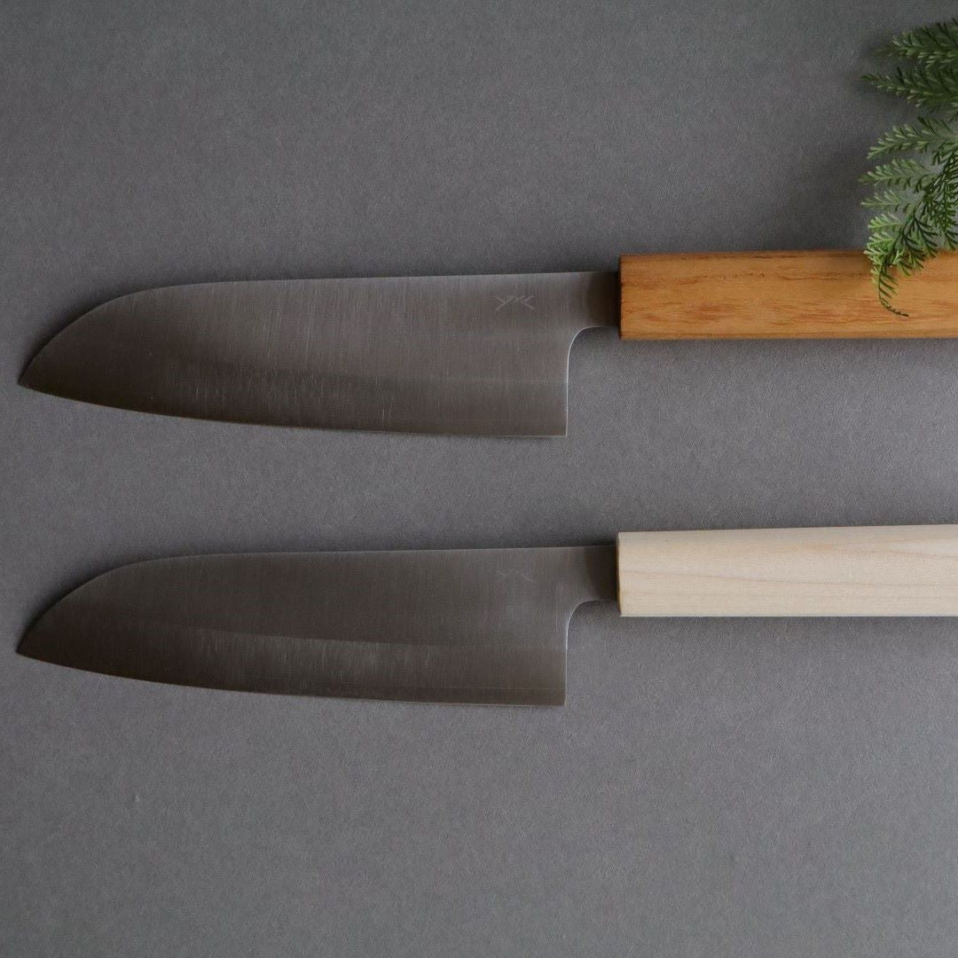 Two Japanese knives with sleek stainless steel blades and wooden handles. The knife at the top has a darker wooden handle, while the one at the bottom has a lighter wooden handle. Both knives are placed on a flat gray surface with a small sprig of green foliage in the upper right corner.