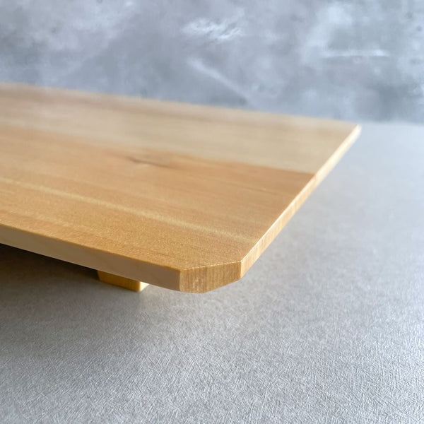 A side view of a light-colored Hinoki wood tray resting on a grey surface, with a textured concrete wall in the background. The tray is slightly elevated, with its smooth, polished surface and subtly rounded edges catching the light, showcasing the fine craftsmanship and natural beauty of the wood.