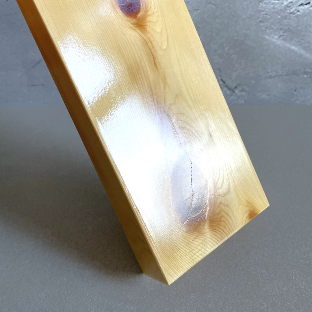 A close-up image of a glossy, varnished pine shelf with a visible wood grain and knots, set at an incline against a grey concrete backdrop. The angle highlights the shelf&