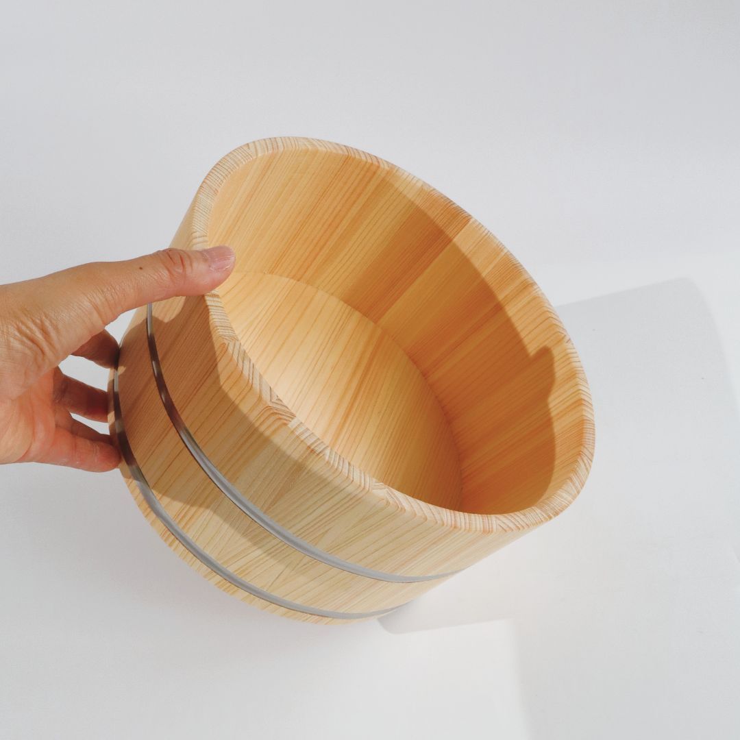 A hand holding a light natural wood bath bucket with metal bands, photographed against a clean white background.