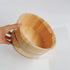 A hand holding a light natural wood bath bucket with metal bands, photographed against a clean white background.