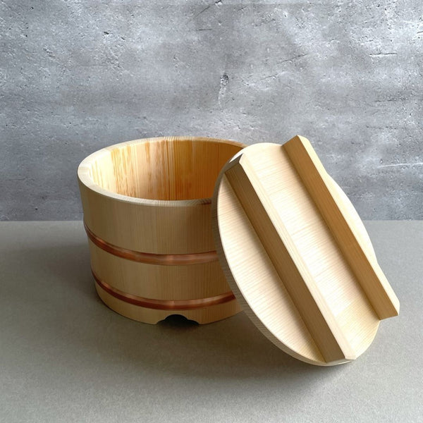 View of a wooden rice bucket with its lid open, positioned on a light brown table in a grey room. The lid is lying on the edge of the bucket.