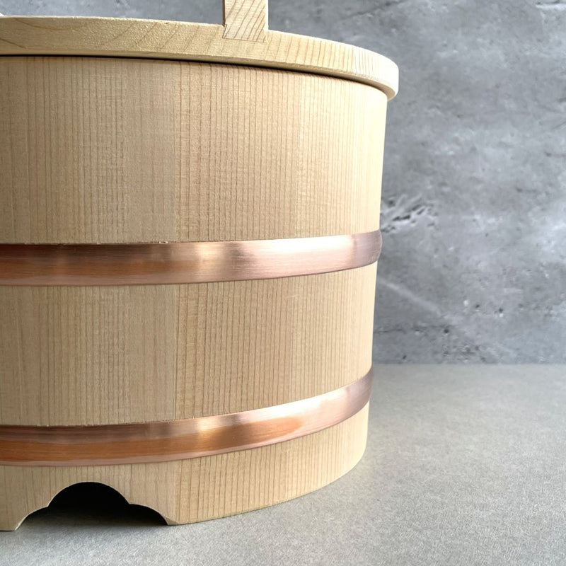 Close-up view of a wooden rice bucket with a copper loop, placed on a light brown table in a grey room.