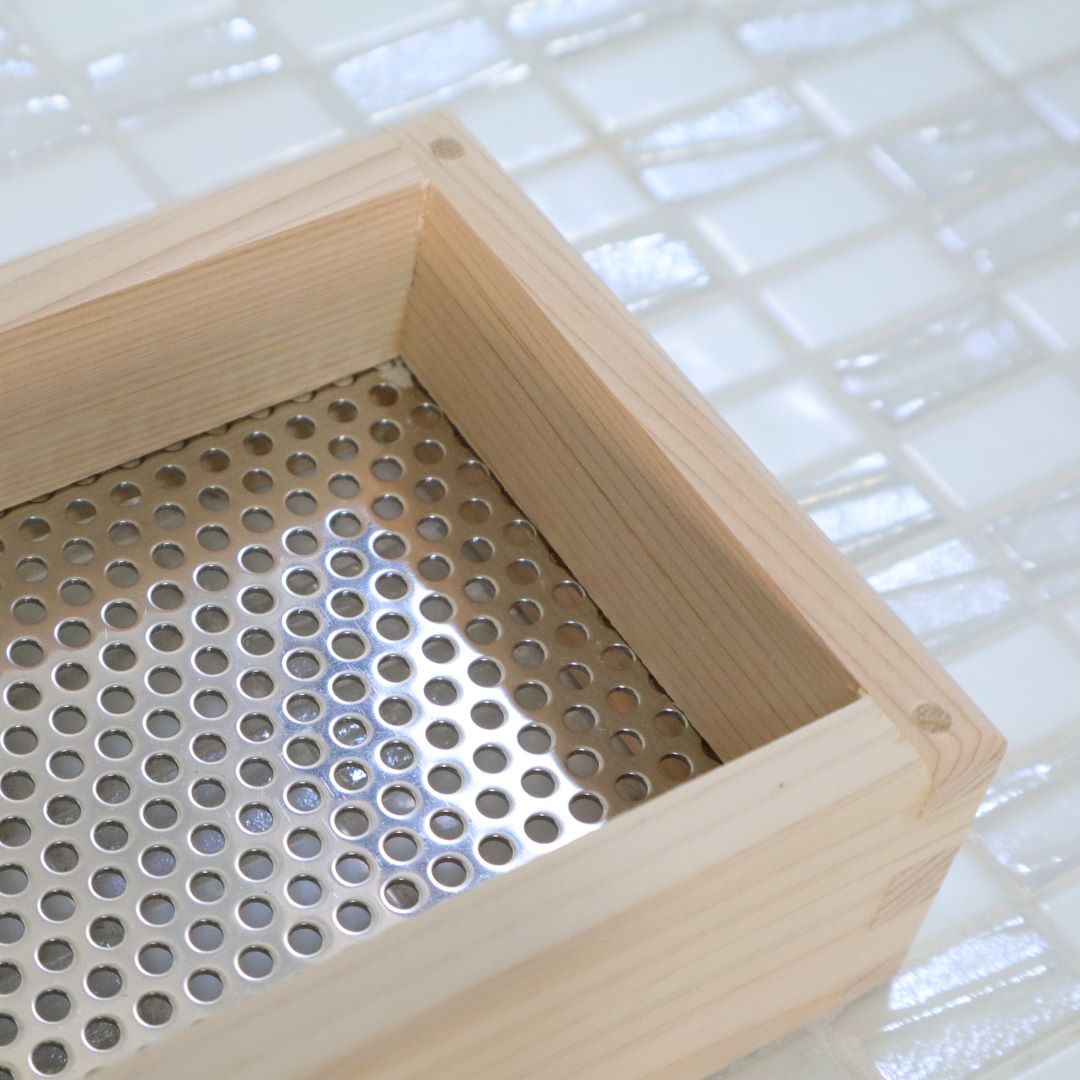 A close-up of the corner of a light hinoki cypress wooden soap dish, showcasing its fine craftsmanship and joinery, set against a tiled bathroom backdrop with a perforated metal soap holder visible within.