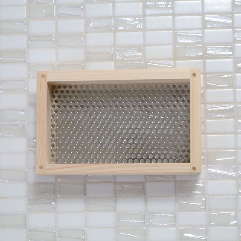  A top-down view of a rectangular hinoki cypress wooden soap dish with a perforated stainless steel panel, resting on a glossy white tiled surface, demonstrating a blend of natural wood textures and modern design.