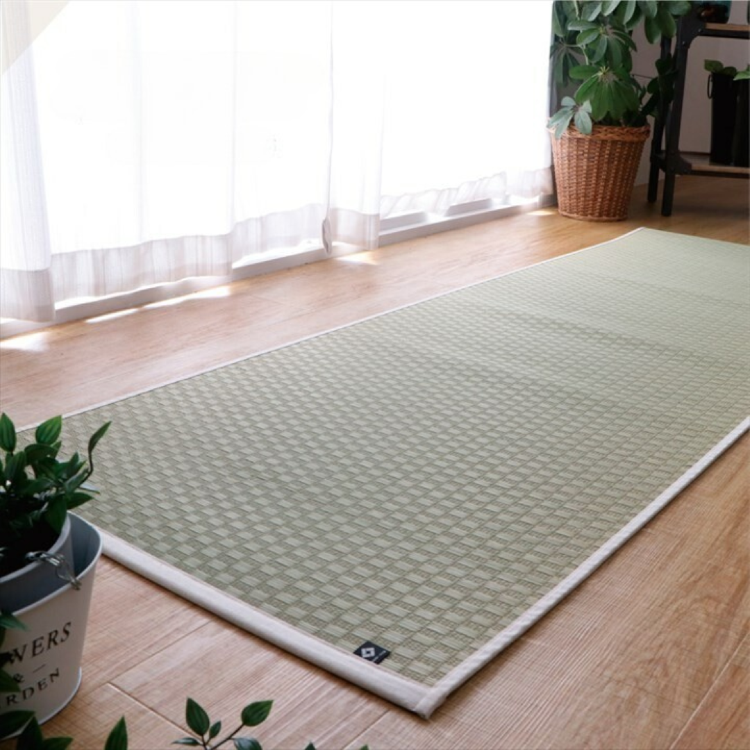 Japanese rush grass tatami mat with a checkered pattern laid on a wooden floor in a bright room with natural light. The mat features a white border and a non-slip backing, surrounded by green plants and minimalist decor.