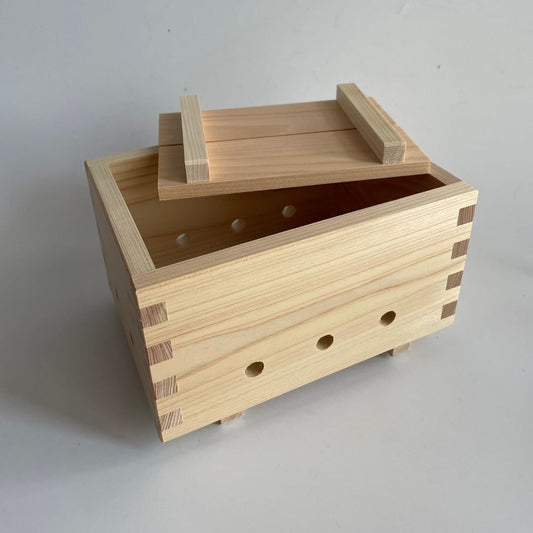 A wooden tofu mold with a lightly open lid, revealing three holes at the front, set in the middle of a grey room.
