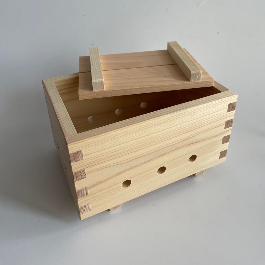 A wooden tofu mold with a lightly open lid, revealing three holes at the front, set in the middle of a grey room.