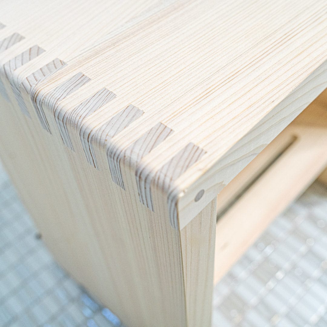 This is a close-up view of a corner of a Japanese square wooden shower stool. The natural texture of the wood is visible, with the grains and knots adding character to the stool's surface. The square shape of the stool is emphasized, and the sturdy construction can be seen in the joint at the corner.