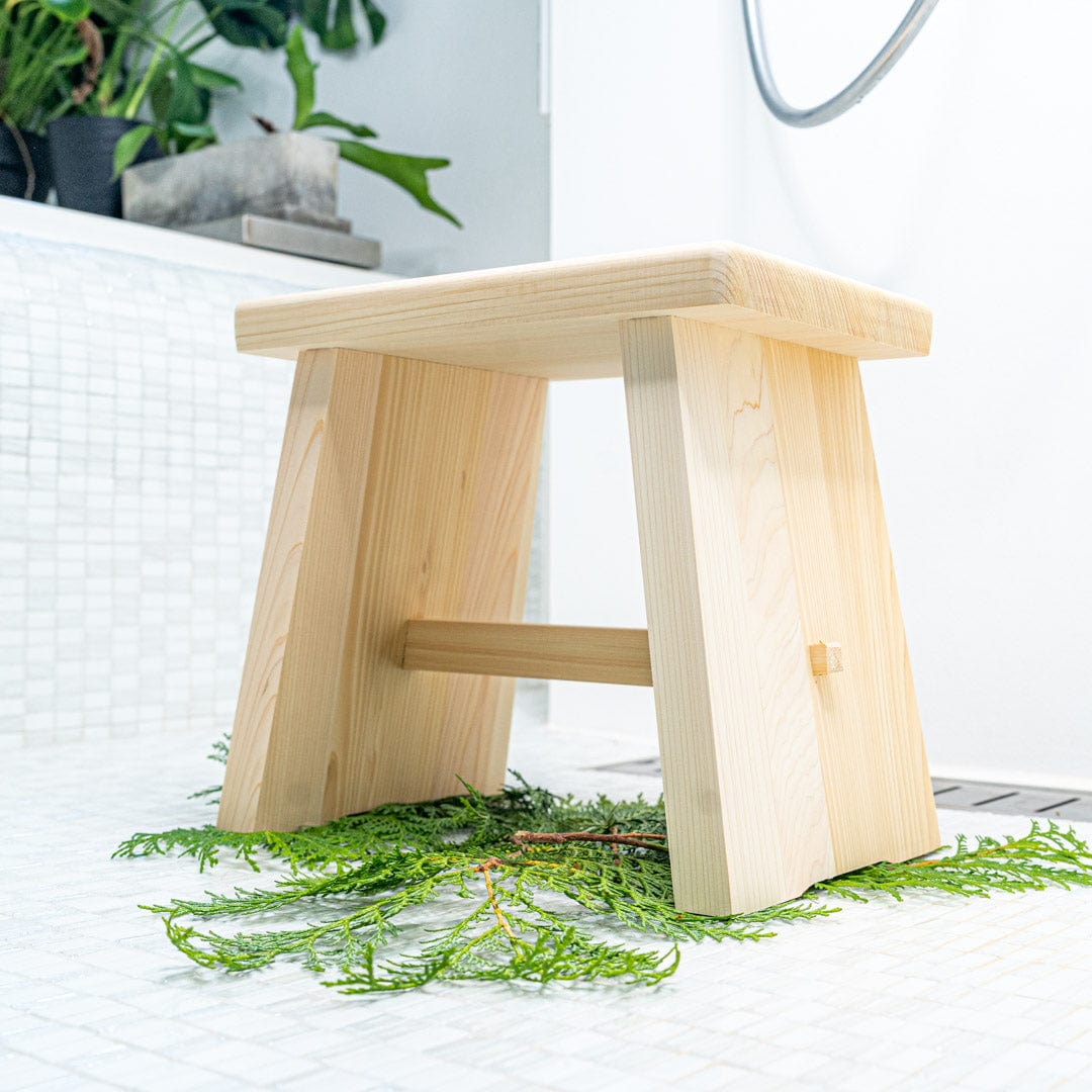A Japanese wooden bath stool is placed in the middle of a bathroom with white faience tiles covering the walls. The stool is positioned next to a bath and there are green plants on the bath, adding a natural touch to the bathroom decor.