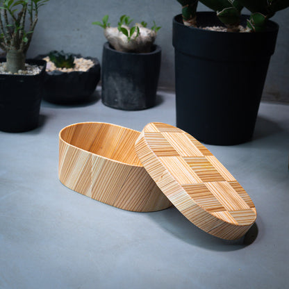 An oval-shaped Japanese cedar wood lunch bento box with an open lid placed on a grey kitchen counter, with green plants in the background.