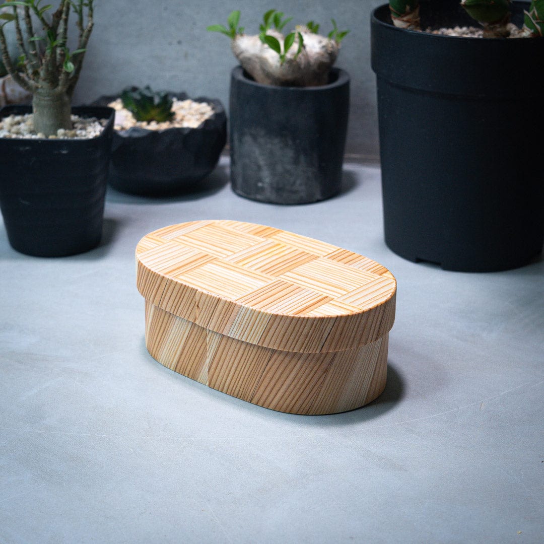  An oval-shaped Japanese cedar wood lunch bento box placed on a grey kitchen counter, with green plants in the background.