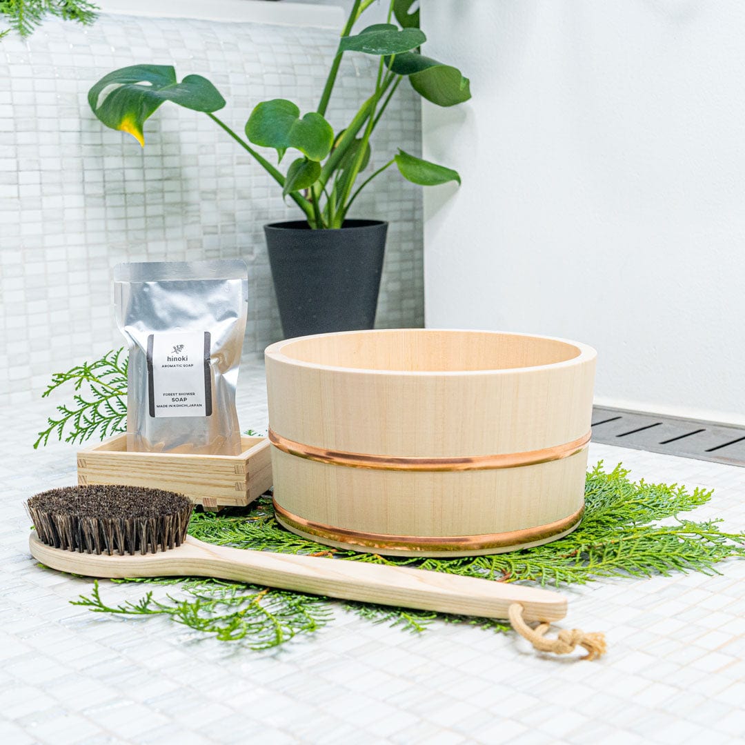 In a white ceramic bathroom, a wooden bath bucket and a wooden body brush with black hair are placed next to a soap dish with soap inside, all positioned in front a green plant.