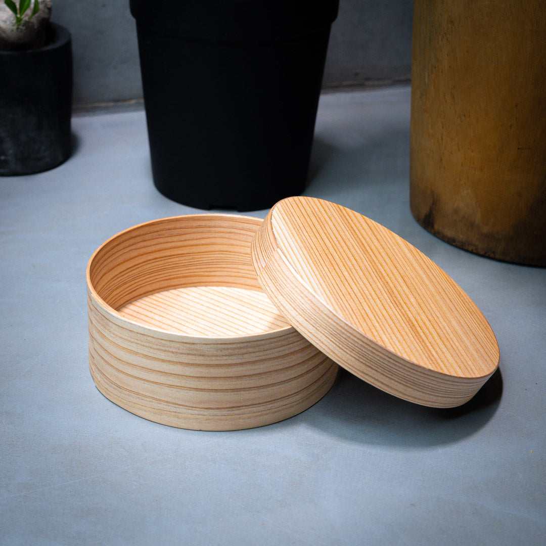 A round Japanese cedar wood lunch bento box is shown in a horizontal view, with its lid open. The bento box is placed on a grey counter, with green plants in the background.