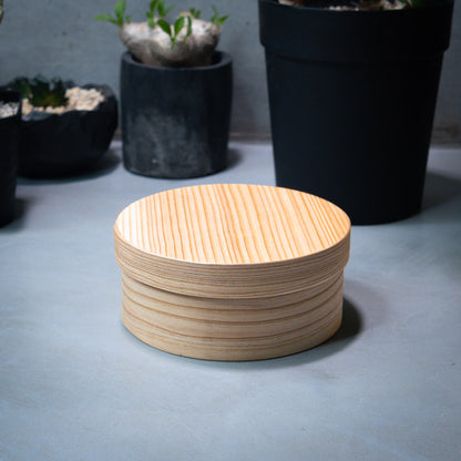 A round Japanese cedar wood lunch bento box with a closed lid is pictured in a horizontal view. The bento box is placed on a grey counter, with green plants in the background.