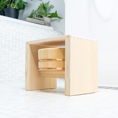 Wooden square stool with a wooden bath bucket in a japanese white bathroom with plants on the background