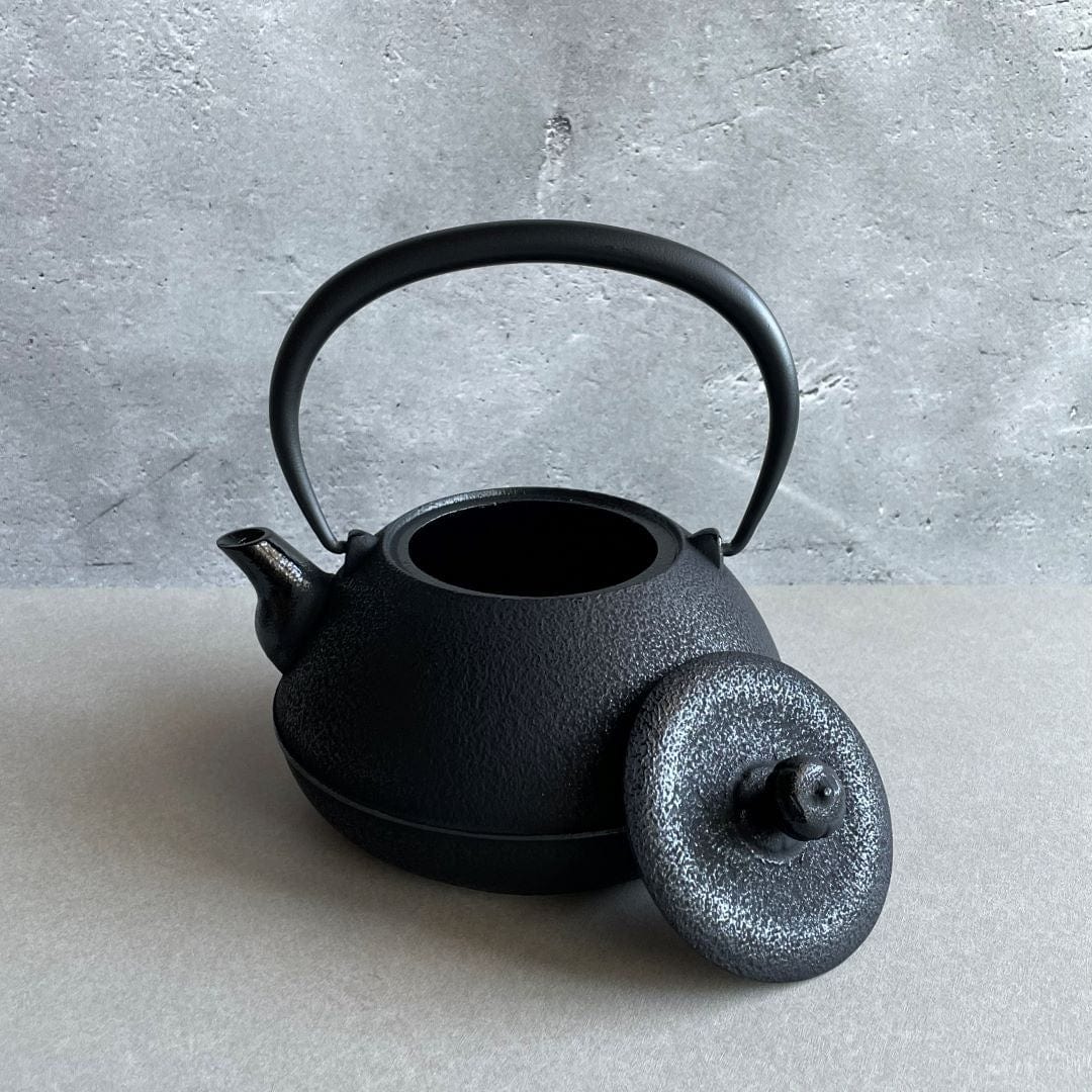 A black cast iron tetsubin kettle with lightly rough surface  on the body, the iron handle has smooth surface and , the kettle is standing in the middle of a space with grey wall and grey surface, the lid is laid on the edge of the kettle.