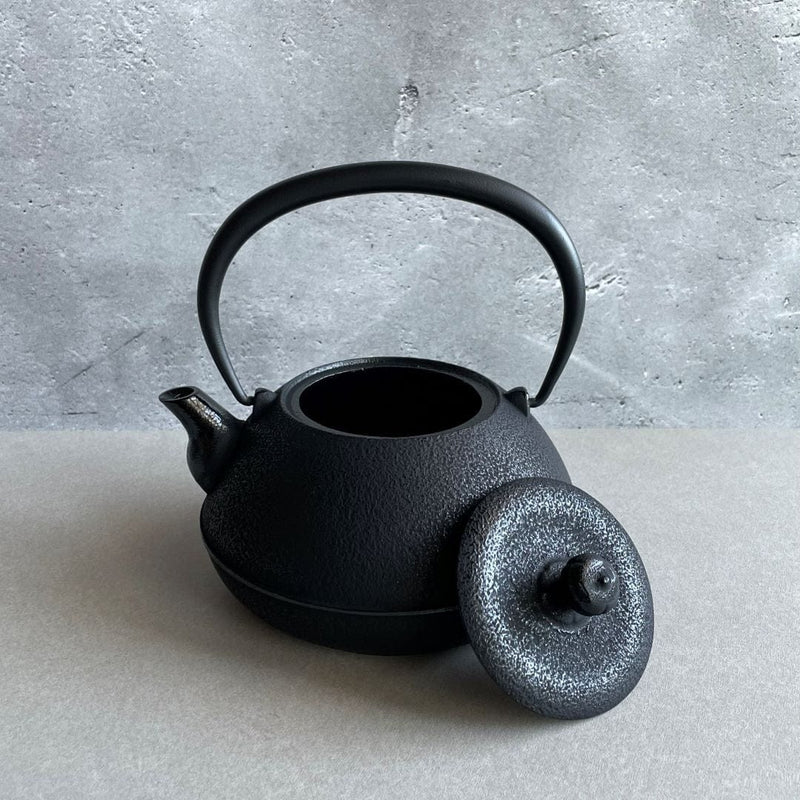 A black cast iron tetsubin kettle with lightly rough surface  on the body, the iron handle has smooth surface and , the kettle is standing in the middle of a space with grey wall and grey surface, the lid is laid on the edge of the kettle.