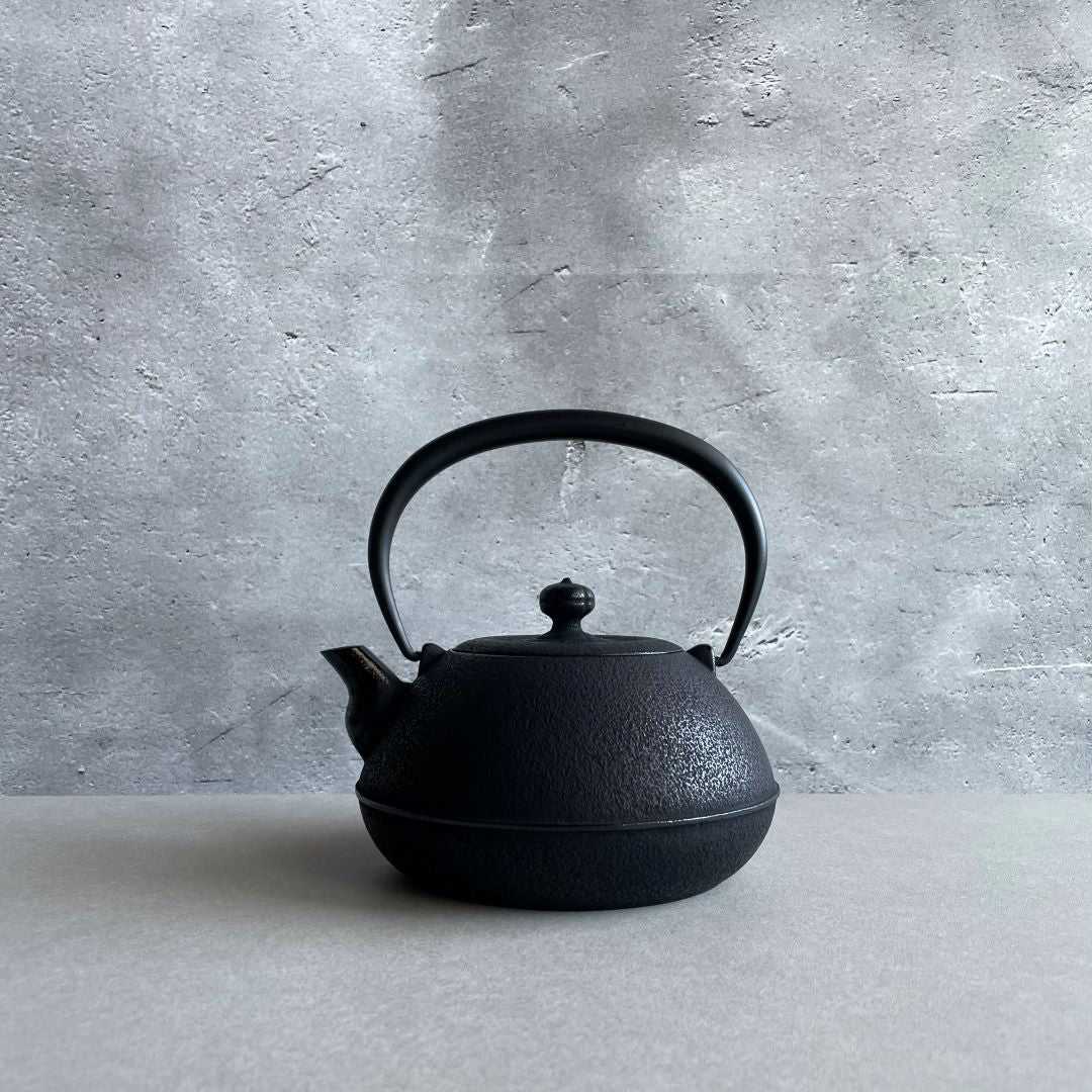 This is a black cast iron Tetsubin kettle with a lightly rough surface on the body. The iron handle has a smooth surface and is standing upright. The kettle is placed in the center of a space with grey walls and a grey surface, seen from a front view.