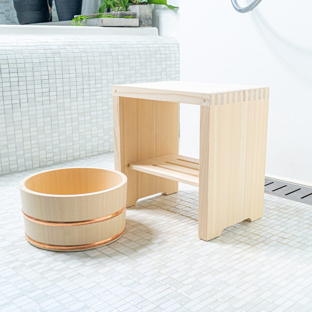 Square bath stool alongside a hinoki wood bucket in a white ceramic bathroom. Green plants enhance the natural atmosphere.