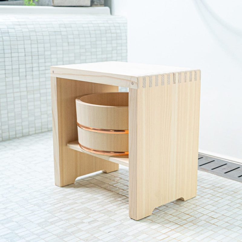 Hinoki wood bucket placed inside a Hinoki wood stool in a white ceramic bathroom. Green plants add a touch of nature to the ambiance.