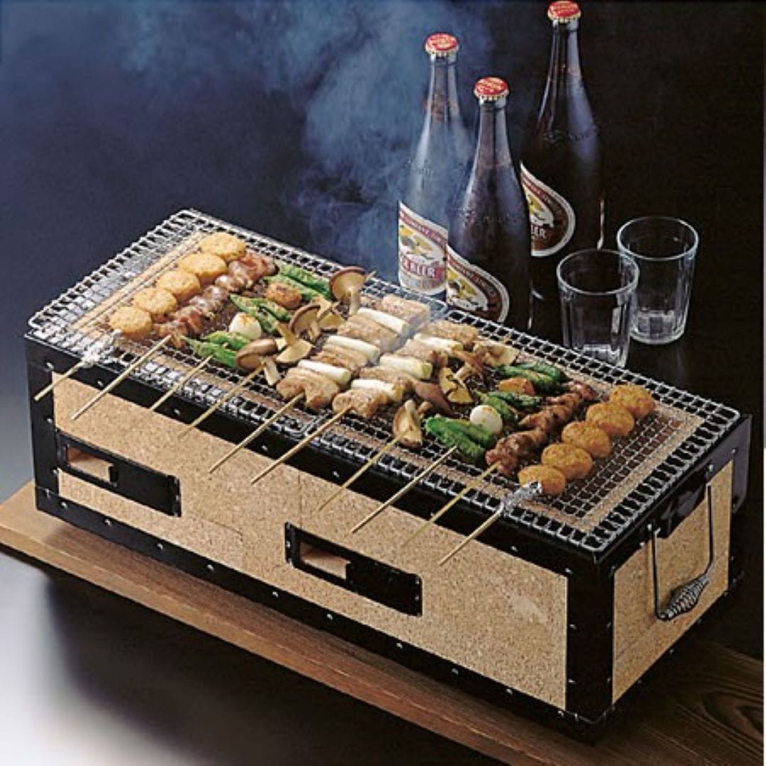 A rectangular-shaped Japanese hibachi charcoal grill is viewed from one corner in a black room. On the iron grill on top of the brown ceramic BBQ, there are yakitori meat sticks and vegetables being cooked. Beer bottles can be seen behind the hibachi.