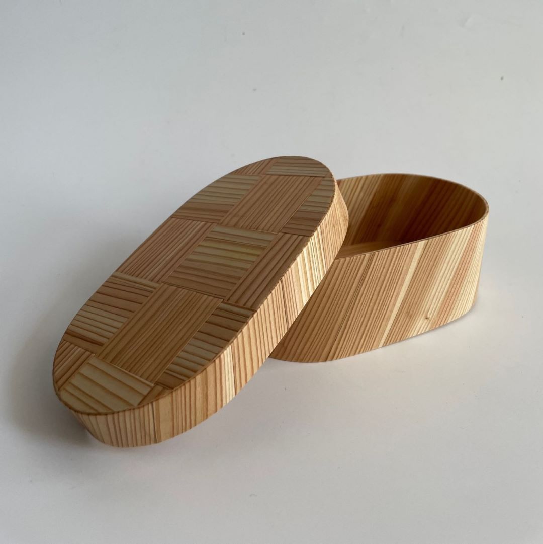 An oval-shaped Japanese cedar wood lunch bento box with an open lid placed on a white surface.