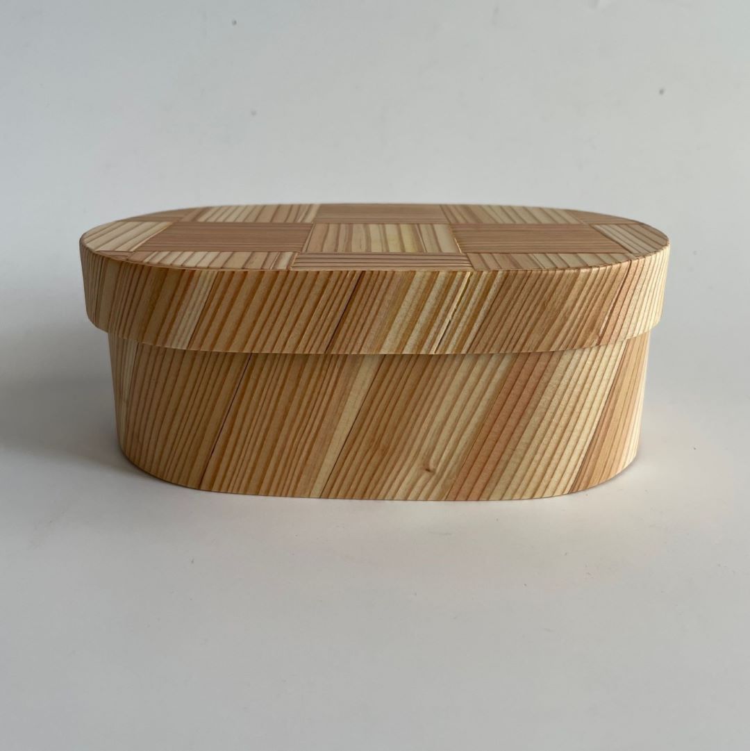 A Japanese cedar wood lunch bento box with an oval shape is shown in a horizontal view. The lid of the bento box is closed, and the photo is taken in a white room.