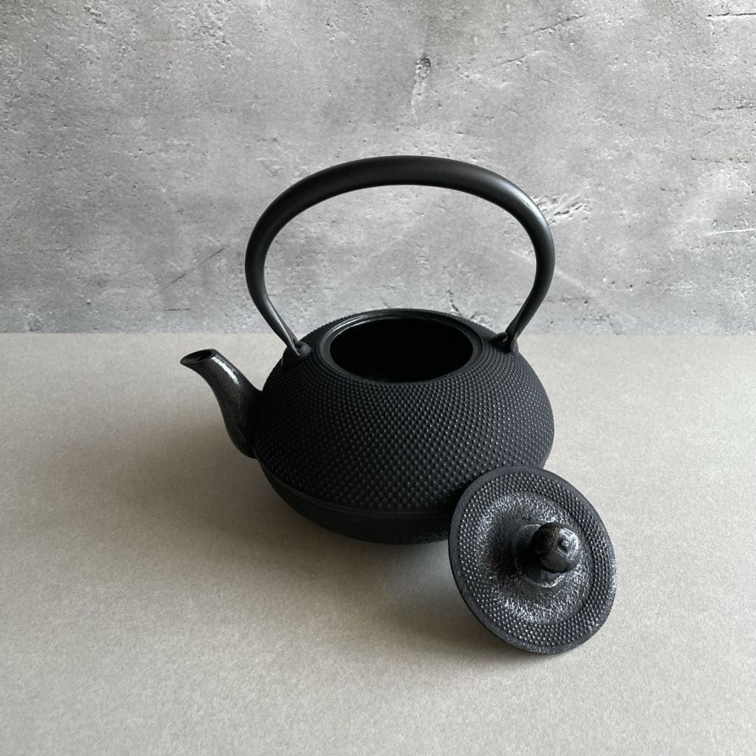 A black cast iron tetsubin kettle with lightly rough surface on the body, the iron handle has smooth surface and, the kettle is standing in the middle of a space with grey wall and grey surface, the lid is laid on the edge of the kettle.