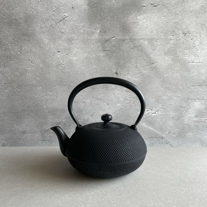 This is a black cast iron Tetsubin kettle with a lightly rough surface on the body. The iron handle has a smooth surface and is standing upright. The kettle is placed in the center of a space with grey walls and a grey surface, seen from a front view.