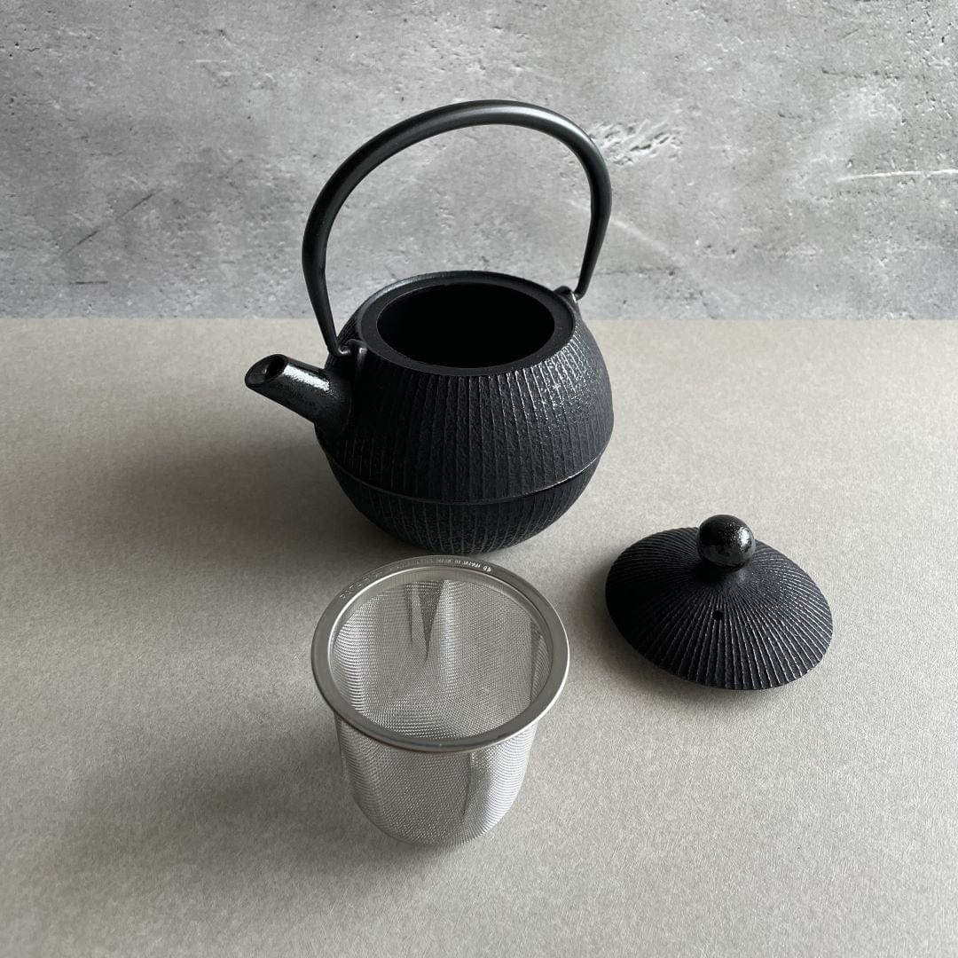 An open round black cast iron tetsubin teapot with vertical strip pattern on the body and a smooth iron handle, standing on a grey surface in a grey room. The lid is off and resting on a brown table beside the teapot, the stainless steel infuser inside is also outside next to the teapot.
