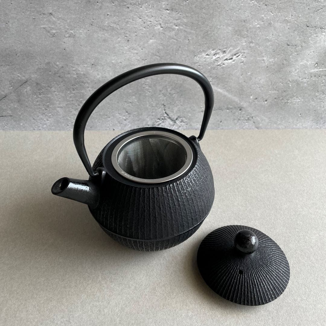 An open round black cast iron tetsubin teapot with vertical strip pattern on the body and a smooth iron handle, standing on a grey surface in a grey room. The lid is off and resting on a brown table beside the teapot, revealing the stainless steel infuser inside.