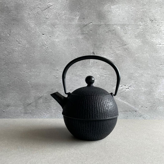 This is a front view of a black cast iron Tetsubin teapot with a lightly rough surface on the body. The teapot has a round shape with vertical stripes all around the surface. The iron handle has a smooth surface and the teapot is standing in the middle of a space with a grey wall and grey surface.