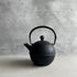 This is a front view of a black cast iron Tetsubin teapot with a lightly rough surface on the body. The teapot has a round shape with vertical stripes all around the surface. The iron handle has a smooth surface and the teapot is standing in the middle of a space with a grey wall and grey surface.