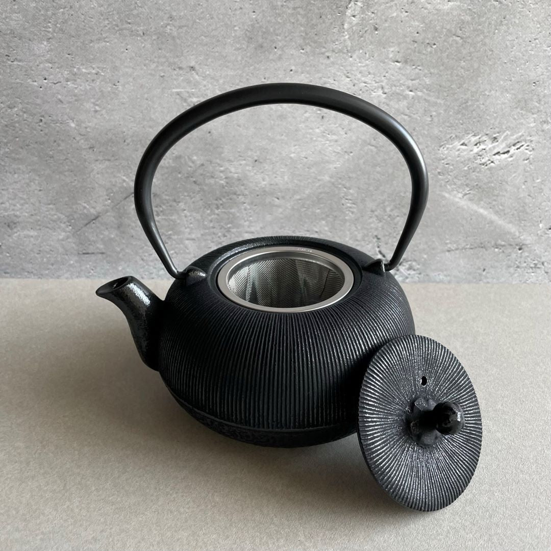 This is a black cast iron Tetsubin teapot with a vertical stripe pattern on the body. The cast iron handle has a smooth surface and is standing upright. The teapot is placed in the center of a space with grey walls and a grey surface. The lid is open and laid on the edge of the teapot, revealing a stainless steel infuser inside.