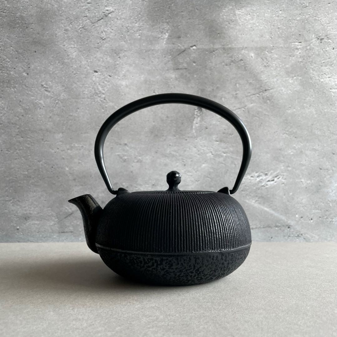This is a black cast iron Tetsubin teapot with a vertical stripe pattern on the body and a rough surface underneath. The iron handle has a smooth surface and is standing upright. The teapot is placed in the center of a space with grey walls and a grey surface.