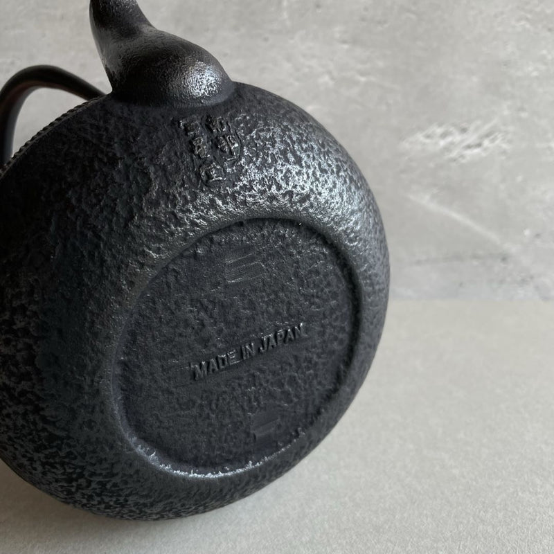 This is the underside of a black cast iron Tetsubin teapot with a rough surface. The words "Made in Japan" are written on the surface. The teapot is laid on its body, revealing the rough texture of the underside.