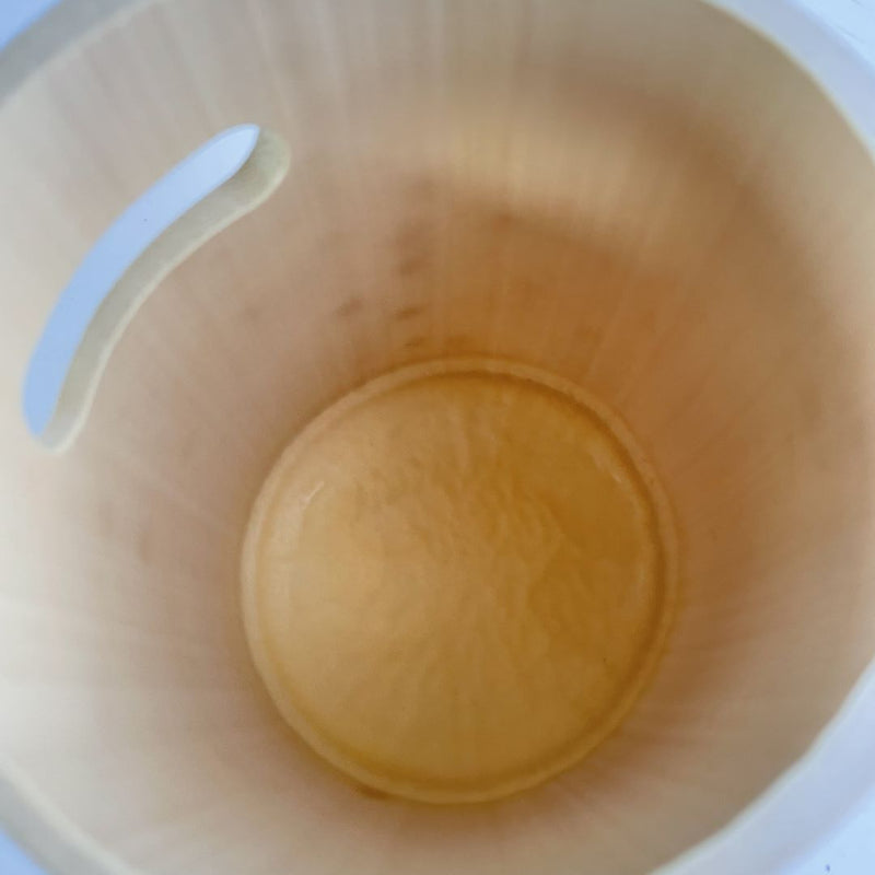 A top-down view inside a light wooden bucket, showing a small amount of a yellowish liquid pooled at the bottom. The interior of the bucket displays a smooth, natural wood grain pattern, and the rim has a thin, white handle attached on one side. The focus is soft, with the central part of the image slightly blurred, emphasizing the presence of the liquid.