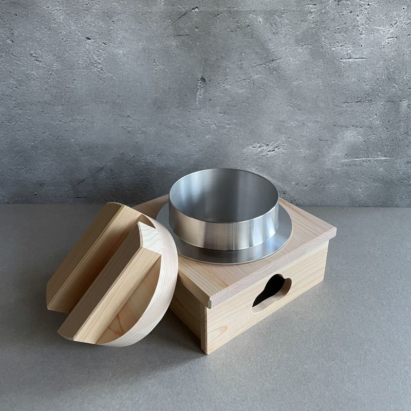 A wooden rice cooker sits in the center of a grey background. Beside it, a wooden lid rests to the side of an aluminum pot, which is nestled inside a wooden stand.