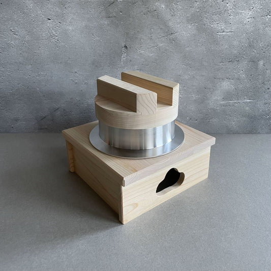A wooden rice cooker sits in the center of a plain grey background. The rice cooker consists of an aluminum pot with a wooden lid resting on top, and the pot is nestled inside a wooden stand. The minimalist design emphasizes the natural beauty of the wood and metal materials.