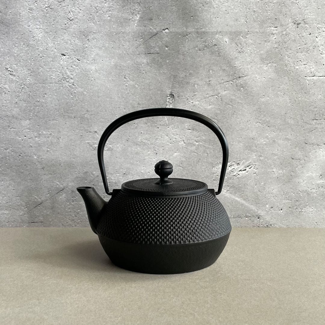 This is a black cast iron Tetsubin kettle with a lightly rough surface on the body. The iron handle has a smooth surface and is standing upright. The kettle is placed in the center of a space with grey walls and a grey surface, seen from a front view.