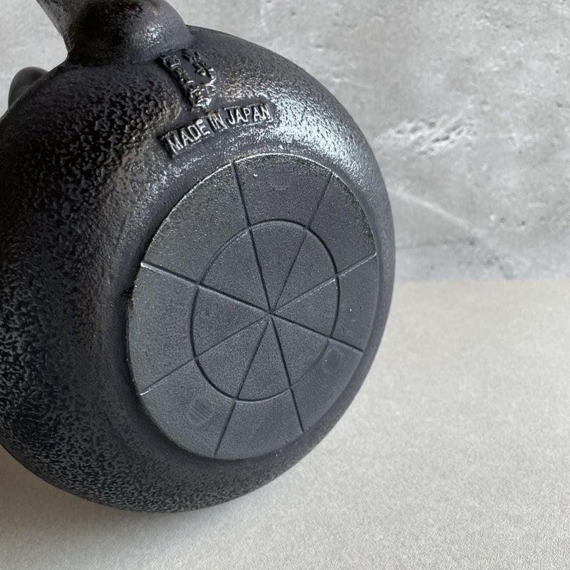 This is the underside of a blue cast iron Tetsubin teapot with a rough surface. The words "Made in Japan" are written on the surface. The teapot is laid on its body, revealing the rough texture of the underside.