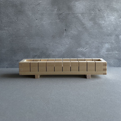 Front view of a rectangular wooden sushi mold resting on a gray surface with a gray wall in the background.
