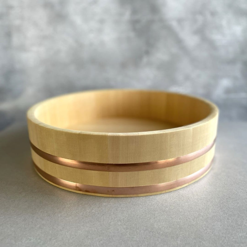 Front view of a brown wooden Hangiri sushi bucket with two copper rings around the rim, placed on a grey surface against a grey background.