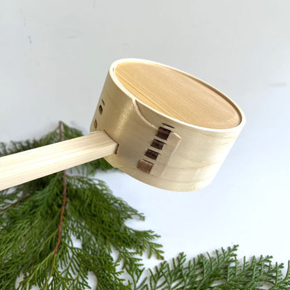Back view of the top section of a wooden sauna ladle resting on a pine branch, with a gray room in the background.
