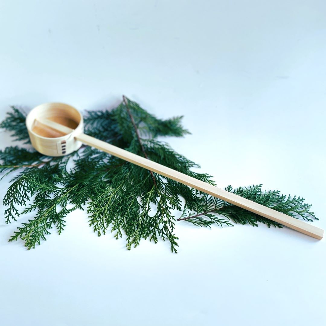 A wooden sauna ladle made of Hinoki wood rests on a pine branch in a minimalist grey room, providing a natural and calming aesthetic.