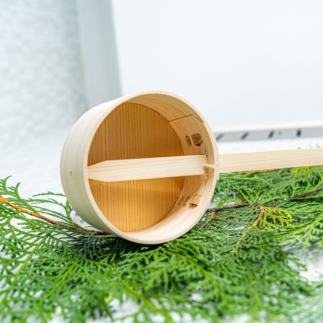 Top view of a wooden sauna ladle resting on a pine branch against a grey background.