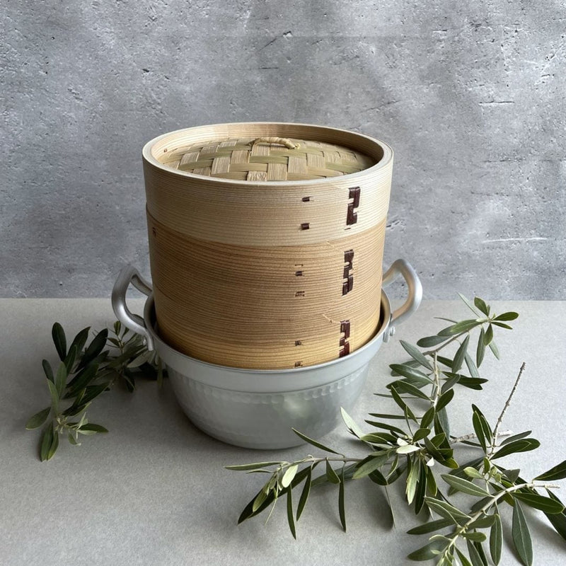 A simple composition with a grey wall in the background and a grey table in the foreground. In the center, a brown bamboo steamer basket is placed, surrounded by two olive branches on each side. The neutral colors and natural elements create a calming and organic vibe.