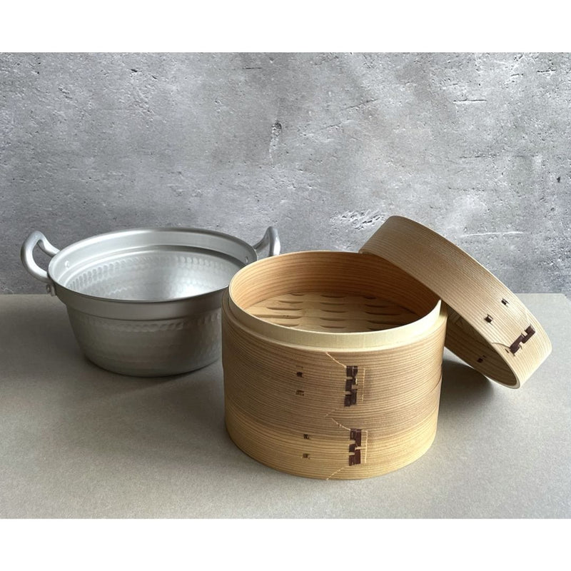 A functional kitchen scene with a plain grey wall in the background and a grey table in the foreground. In the center of the table, a brown bamboo steamer basket with its lid open is displayed, ready to serve its contents. Next to it, an aluminum pot is placed, suggesting a cooking process. The mix of natural and man-made materials creates an interesting contrast, while the muted color palette gives a modern and elegant feel to the overall composition.