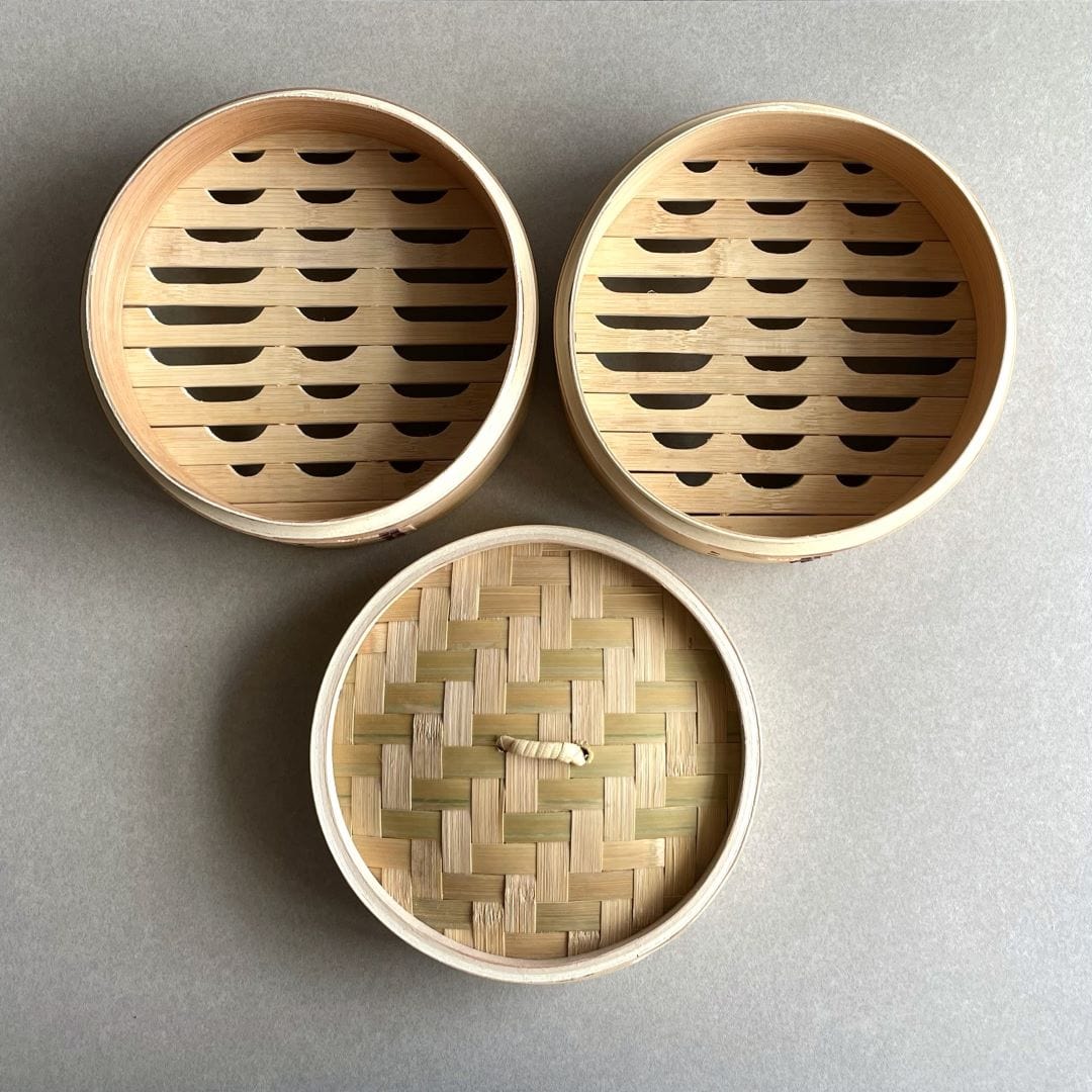 A simple yet intriguing setup with a plain grey tabletop as the backdrop. On top of the table, two brown bamboo steamer baskets are placed, one on top of the other, creating a harmonious stack. A lid is included, indicating the possibility of keeping the steamed food warm before serving. The natural texture of the bamboo adds warmth and depth to the composition, while the neutral colors make it easy to fit into any decor style.